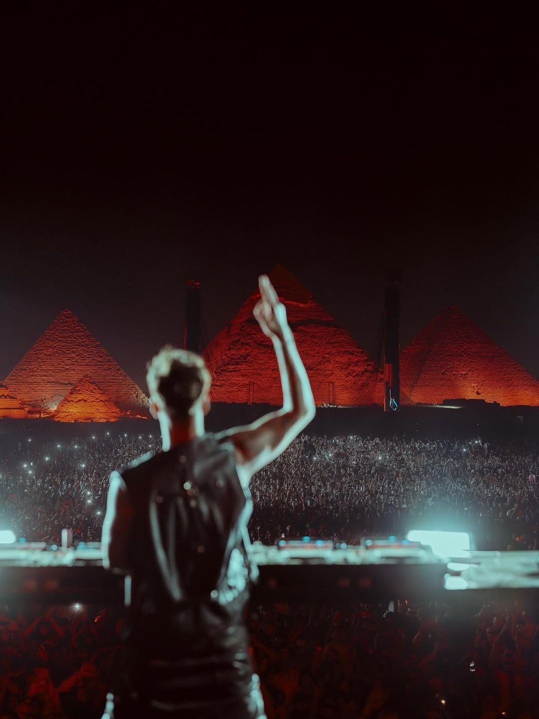 A DJ facing a crowd of thousands, with the pyramids of Giza lit up red behind